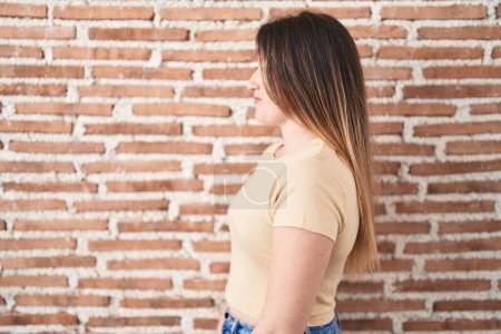 Photo for Young brunette woman standing over bricks wall looking to side, relax profile pose with natural face and confident smile. - Royalty Free Image
