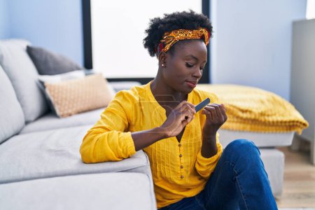 Téléchargez les photos : Jeune femme afro-américaine utilisant des ongles lime assis sur le sol à la maison - en image libre de droit