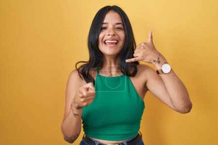 Photo for Brunette woman standing over yellow background smiling doing talking on the telephone gesture and pointing to you. call me. - Royalty Free Image