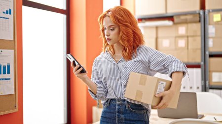 Téléchargez les photos : Jeune rousse femme commerce électronique travailleur d'entreprise tenant paquet à l'aide d'un smartphone au bureau - en image libre de droit