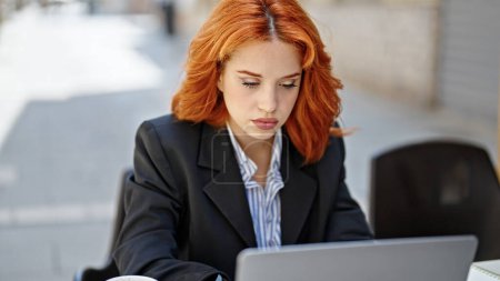 Téléchargez les photos : Jeune rousse femme d'affaires utilisant un ordinateur portable avec une expression détendue sur la terrasse du café - en image libre de droit