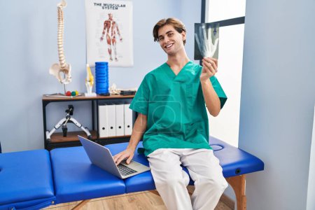 Foto de Joven hombre caucásico fisioterapeuta buscando rayos X utilizando ordenador portátil en la clínica de rehabilitación - Imagen libre de derechos