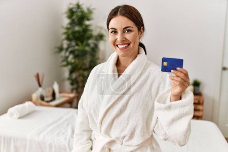 Téléchargez les photos : Jeune belle femme hispanique portant peignoir tenant carte de crédit au salon de beauté - en image libre de droit