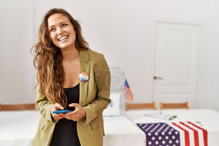 Téléchargez les photos : Jeune belle femme hispanique président de table électorale en utilisant smartphone au collège électoral - en image libre de droit