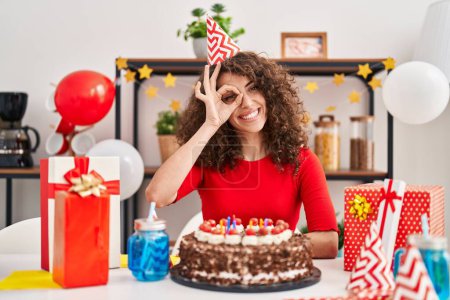 Téléchargez les photos : Femme hispanique avec les cheveux bouclés célébrant anniversaire tenant grand gâteau au chocolat souriant heureux faire ok signe avec la main sur les yeux regardant à travers les doigts - en image libre de droit