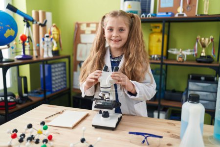 Téléchargez les photos : Adorable fille blonde étudiante souriante confiante en utilisant un microscope dans une salle de classe de laboratoire - en image libre de droit