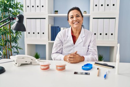 Téléchargez les photos : Dentiste hispanique mature femme travaillant à la clinique regardant positif et heureux debout et souriant avec un sourire confiant montrant les dents - en image libre de droit