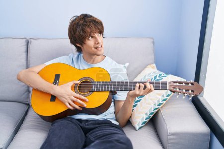 Photo for Young blond man playing classical guitar sitting on sofa at home - Royalty Free Image