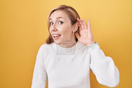 Foto de Mujer caucásica joven con suéter blanco sobre fondo amarillo sonriendo con la mano sobre la oreja escuchando un rumor o chismes. concepto de sordera. - Imagen libre de derechos