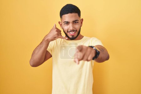 Téléchargez les photos : Jeune homme hispanique debout sur fond jaune souriant faisant parler au téléphone geste et vous pointant du doigt. Appelle-moi. - en image libre de droit