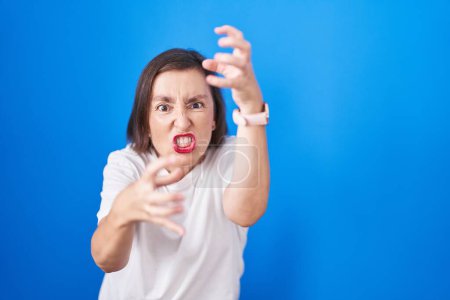 Photo for Middle age hispanic woman standing over blue background shouting frustrated with rage, hands trying to strangle, yelling mad - Royalty Free Image