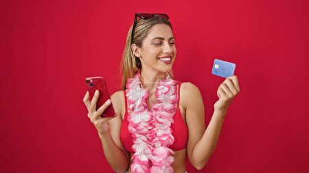 Téléchargez les photos : Jeune femme blonde portant des achats de bikini avec smartphone et carte de crédit sur fond rouge isolé - en image libre de droit