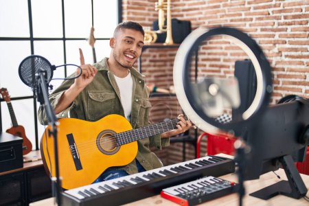 Foto de Joven hombre hispano haciendo tutorial de guitarra en línea sonriendo feliz señalando con la mano y el dedo a un lado - Imagen libre de derechos