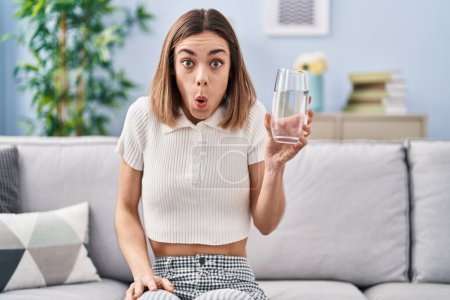 Foto de Mujer hispana bebiendo vaso de agua asustada y asombrada con la boca abierta para sorpresa, cara de incredulidad - Imagen libre de derechos