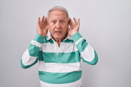 Foto de Hombre mayor con el pelo gris de pie sobre fondo blanco tratando de escuchar ambas manos en el gesto de la oreja, curioso por chismes. problema auditivo, sordo - Imagen libre de derechos
