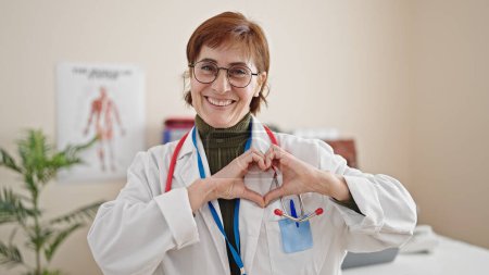 Téléchargez les photos : Femme hispanique mature médecin souriant faisant geste cardiaque avec les mains à la clinique - en image libre de droit