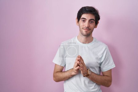 Foto de Joven hombre hispano de pie sobre el fondo rosa manos juntas y los dedos cruzados sonriente relajado y alegre. éxito y optimismo - Imagen libre de derechos