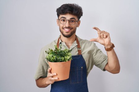Téléchargez les photos : Homme arabe avec barbe tenant pot de plante verte geste souriant et confiant avec la main faisant signe de petite taille avec les doigts à la recherche et la caméra. concept de mesure. - en image libre de droit