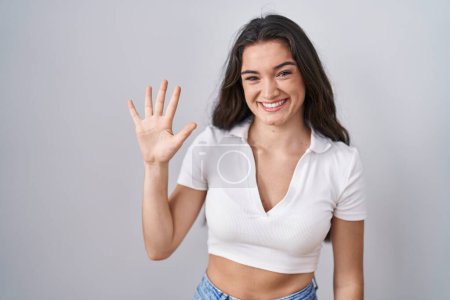 Foto de Joven adolescente de pie sobre fondo blanco mostrando y apuntando hacia arriba con los dedos número cinco mientras sonríe confiado y feliz. - Imagen libre de derechos