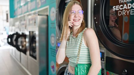 Téléchargez les photos : Jeune femme blonde parlant sur smartphone attendant la machine à laver à la buanderie - en image libre de droit