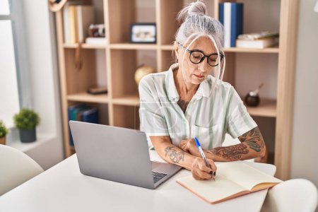 Photo for Middle age grey-haired woman writing on notebook using laptop at home - Royalty Free Image