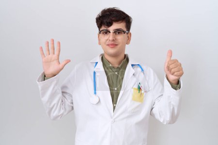 Foto de Joven hombre no binario vistiendo uniforme médico y estetoscopio mostrando y señalando con los dedos número seis mientras sonríe confiado y feliz. - Imagen libre de derechos