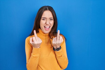 Foto de Young brunette woman standing over blue background showing middle finger doing fuck you bad expression, provocation and rude attitude. screaming excited - Imagen libre de derechos