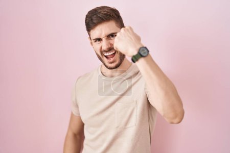 Foto de Hombre hispano con barba de pie sobre fondo rosa enojado y loco levantando el puño frustrado y furioso mientras gritaba con ira. rabia y concepto agresivo. - Imagen libre de derechos