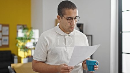 Photo for Young hispanic man business worker drinking coffee reading document at office - Royalty Free Image