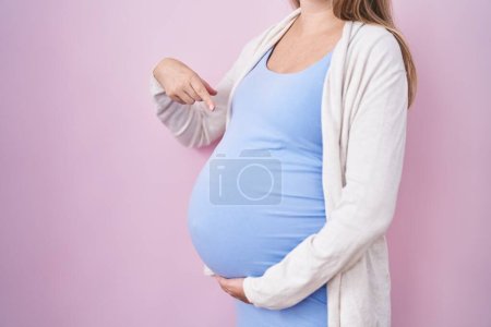 Foto de Mujer embarazada joven esperando un bebé, tocando el vientre embarazada sonriendo feliz señalando con la mano y el dedo - Imagen libre de derechos
