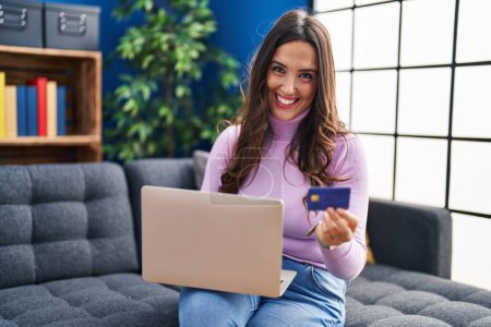 Téléchargez les photos : Jeune femme hispanique utilisant ordinateur portable et carte de crédit assis sur le canapé à la maison - en image libre de droit