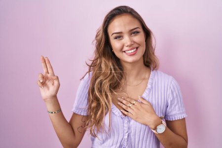 Téléchargez les photos : Jeune femme hispanique debout sur fond rose souriant jurer avec la main sur la poitrine et les doigts vers le haut, faisant un serment de fidélité promesse - en image libre de droit