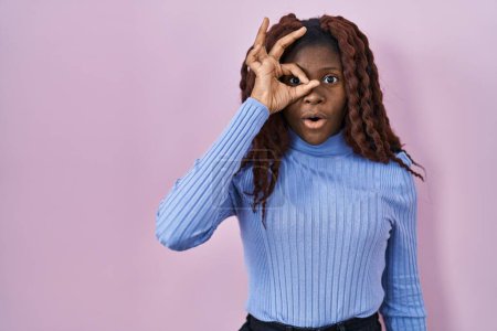 Foto de Mujer africana de pie sobre fondo rosa haciendo gesto ok conmocionado con la cara sorprendida, ojos mirando a través de los dedos. expresión incrédula. - Imagen libre de derechos