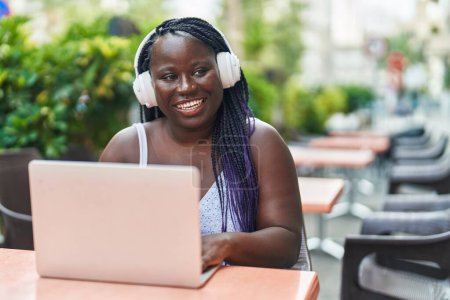 Foto de Mujer afroamericana usando laptop y auriculares sentados en la mesa en la terraza de la cafetería - Imagen libre de derechos