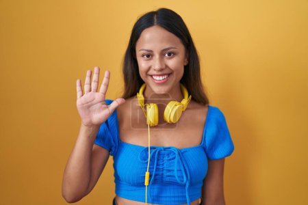 Foto de Mujer joven hispana de pie sobre fondo amarillo mostrando y señalando hacia arriba con los dedos número cinco mientras sonríe confiada y feliz. - Imagen libre de derechos