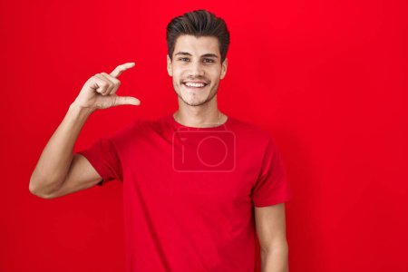 Téléchargez les photos : Young hispanic man standing over red background smiling and confident gesturing with hand doing small size sign with fingers looking and the camera. measure concept. - en image libre de droit