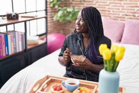 Téléchargez les photos : Femme afro-américaine ayant un petit déjeuner cadeau assis sur le lit dans la chambre - en image libre de droit