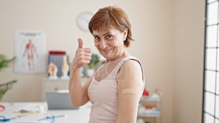 Foto de Mujer hispana madura haciendo pulgares con tirita en el brazo en la clínica - Imagen libre de derechos