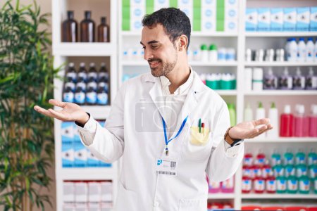 Téléchargez les photos : Beau hispanique homme travaillant à la pharmacie pharmacie souriant montrant les deux mains paumes ouvertes, présentant et la comparaison de la publicité et l'équilibre - en image libre de droit