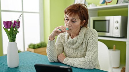 Téléchargez les photos : Mature hispanique femme tenant vape inhalateur assis sur table regarder vidéo sur touchpad à salle à manger - en image libre de droit