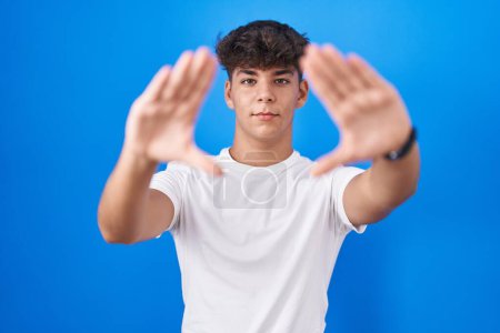 Téléchargez les photos : Adolescent hispanique debout sur fond bleu faisant cadre à l'aide de mains paumes et doigts, perspective de la caméra - en image libre de droit