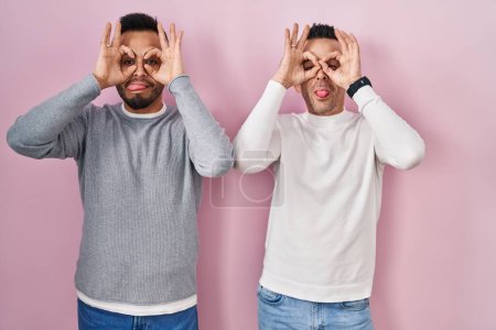 Téléchargez les photos : Couple homosexuel debout sur fond rose faisant un geste correct comme des jumelles sortant la langue, les yeux regardant à travers les doigts. expression folle. - en image libre de droit