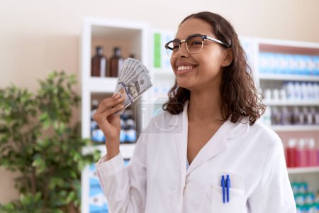 Foto de Joven mujer afroamericana farmacéutica sonriendo confiada sosteniendo dólares en la farmacia - Imagen libre de derechos