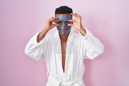 Téléchargez les photos : Jeune homme hispanique portant un masque de beauté et peignoir de bain essayant d'ouvrir les yeux avec les doigts, somnolent et fatigué pour la fatigue matinale - en image libre de droit