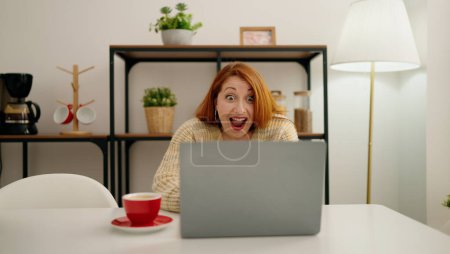 Foto de Joven pelirroja usando laptop con expresión sorpresa en casa - Imagen libre de derechos