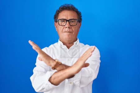 Foto de Hombre hispano de mediana edad de pie sobre fondo azul expresión de rechazo cruzando brazos haciendo signo negativo, cara enojada - Imagen libre de derechos
