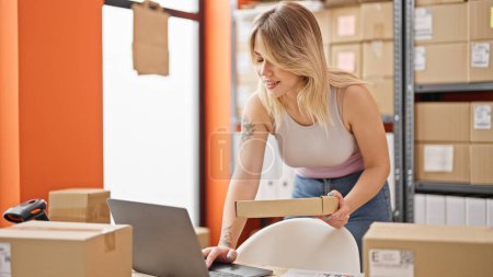 Photo for Young blonde woman ecommerce business worker using laptop holding package at office - Royalty Free Image
