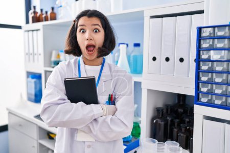 Téléchargez les photos : Jeune femme hispanique travaillant au laboratoire scientifique effrayée et choquée par l'expression surprise et étonnée, la peur et le visage excité. - en image libre de droit