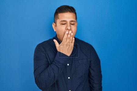 Foto de Joven hispano parado sobre fondo azul aburrido bostezando cansado cubriendo la boca con la mano. inquietud y somnolencia. - Imagen libre de derechos