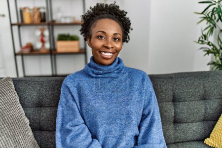 Téléchargez les photos : Femme afro-américaine souriante confiante assise sur un canapé à la maison - en image libre de droit